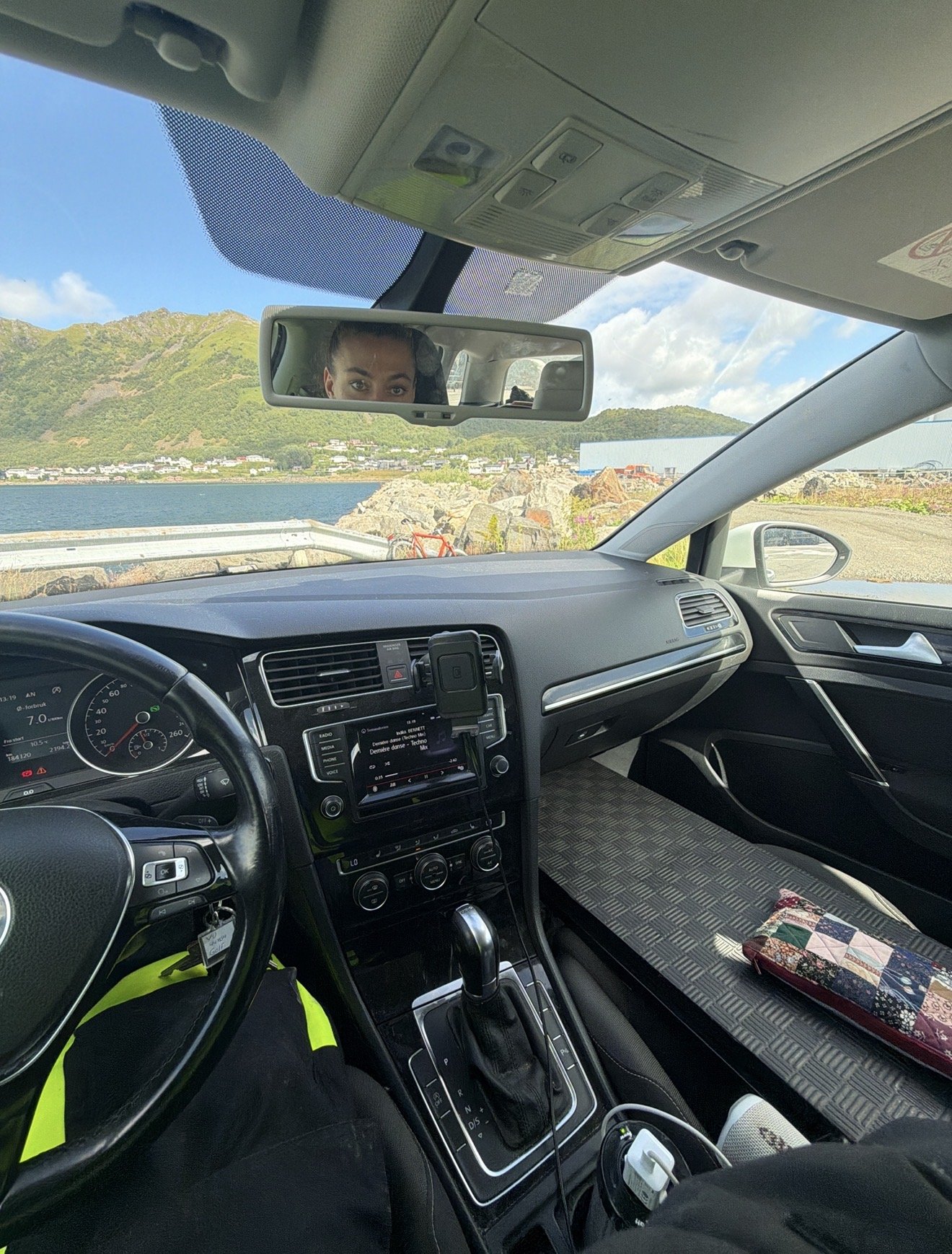 Bilinteriør parkert ved havna, fjell og fjord gjennom frontruta, føreren synlig i sladrespeilet